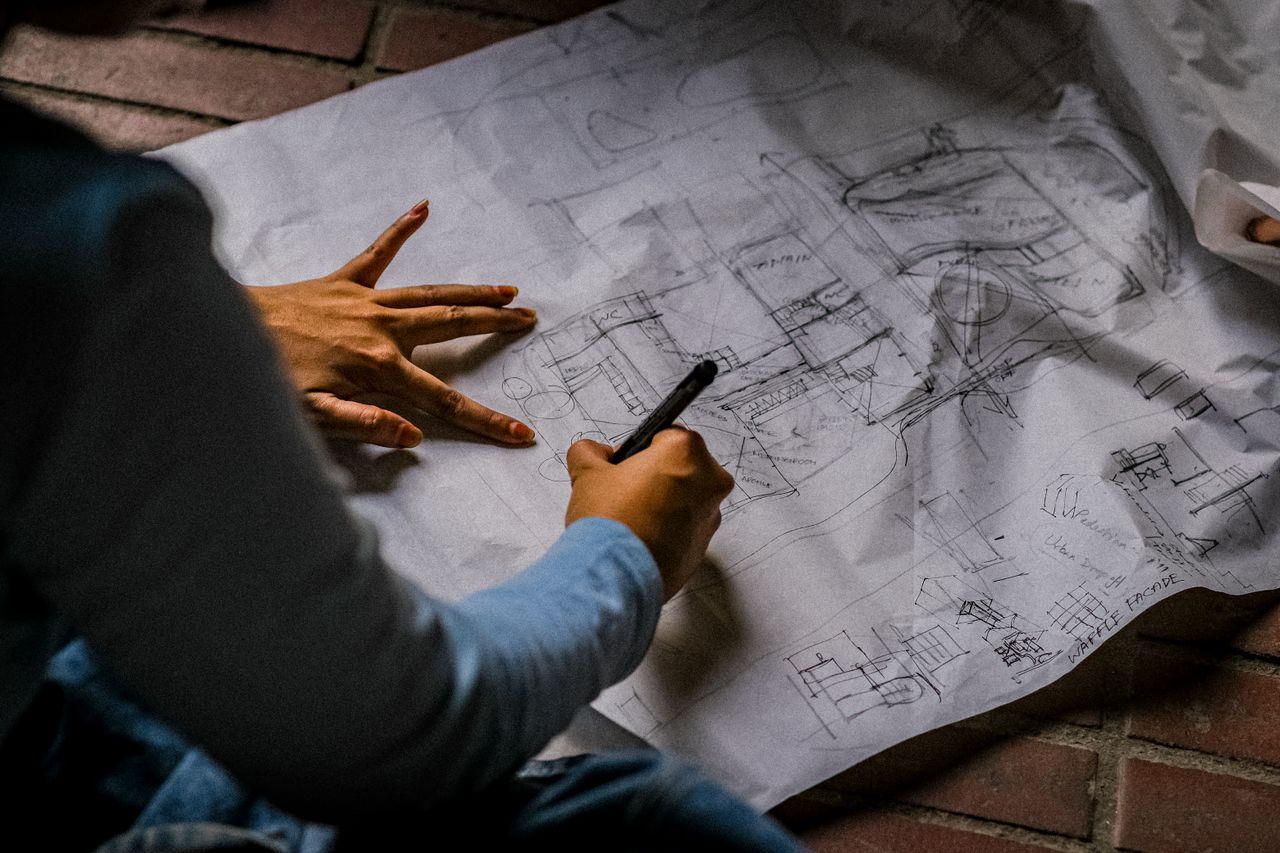 Person sketching architectural design on blueprint paper with pen on brick floor.